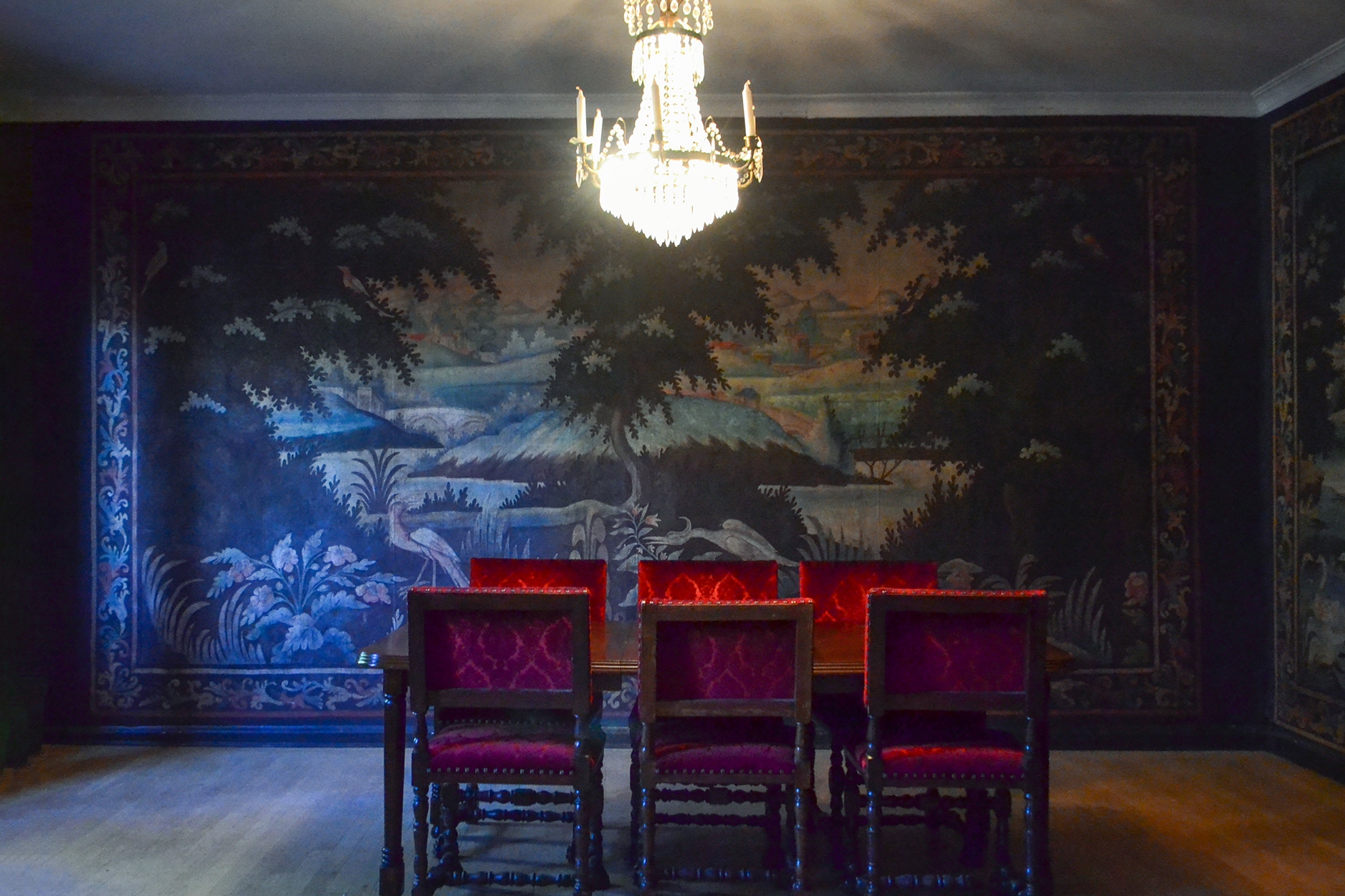 A picture of Italiarummet at Gathenhielmska Huset. Six red chairs, a table and a chandelier. The tapestries on the walls are from Northern Italy and depict an island scene in blues and greens.