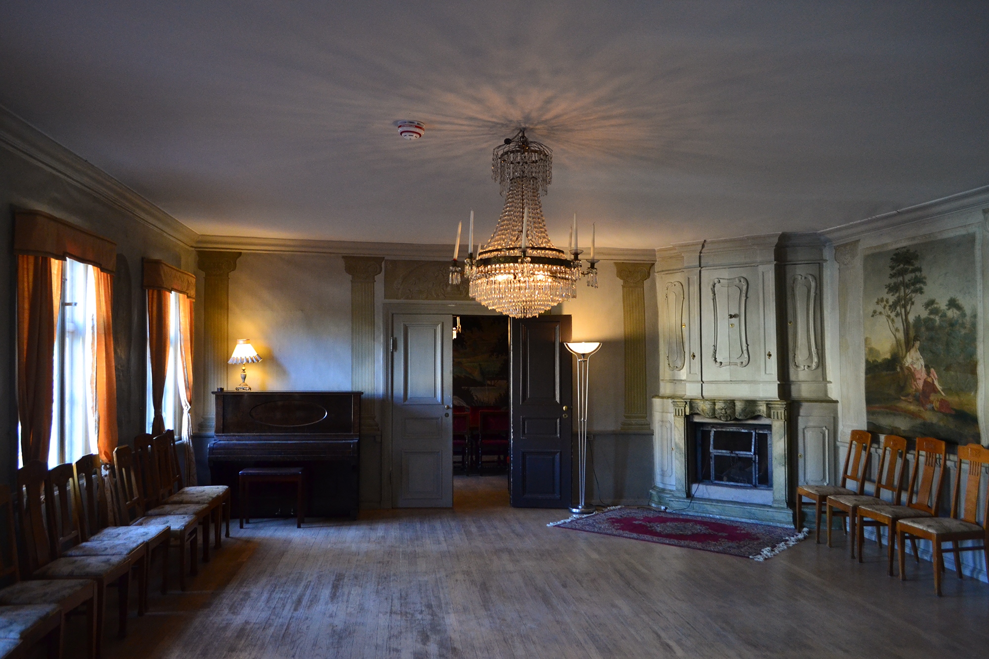 Salongen at Gathenhielmska Huset, with a chandelier, a piano, two lamps, chairs and tapestries on the wall.