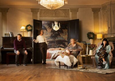 Four actors in a room, one in smart attire sat by a piano, one in smart attire leaning against a door, two in bedclothes sat in armchairs